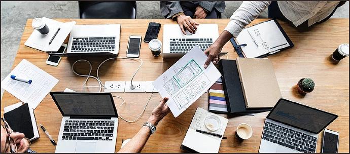 Team collaborating on promotional website and social media page development, with laptops, documents, and design plans spread across the table, focusing on enhancing online presence and visibility for a hospital or clinic