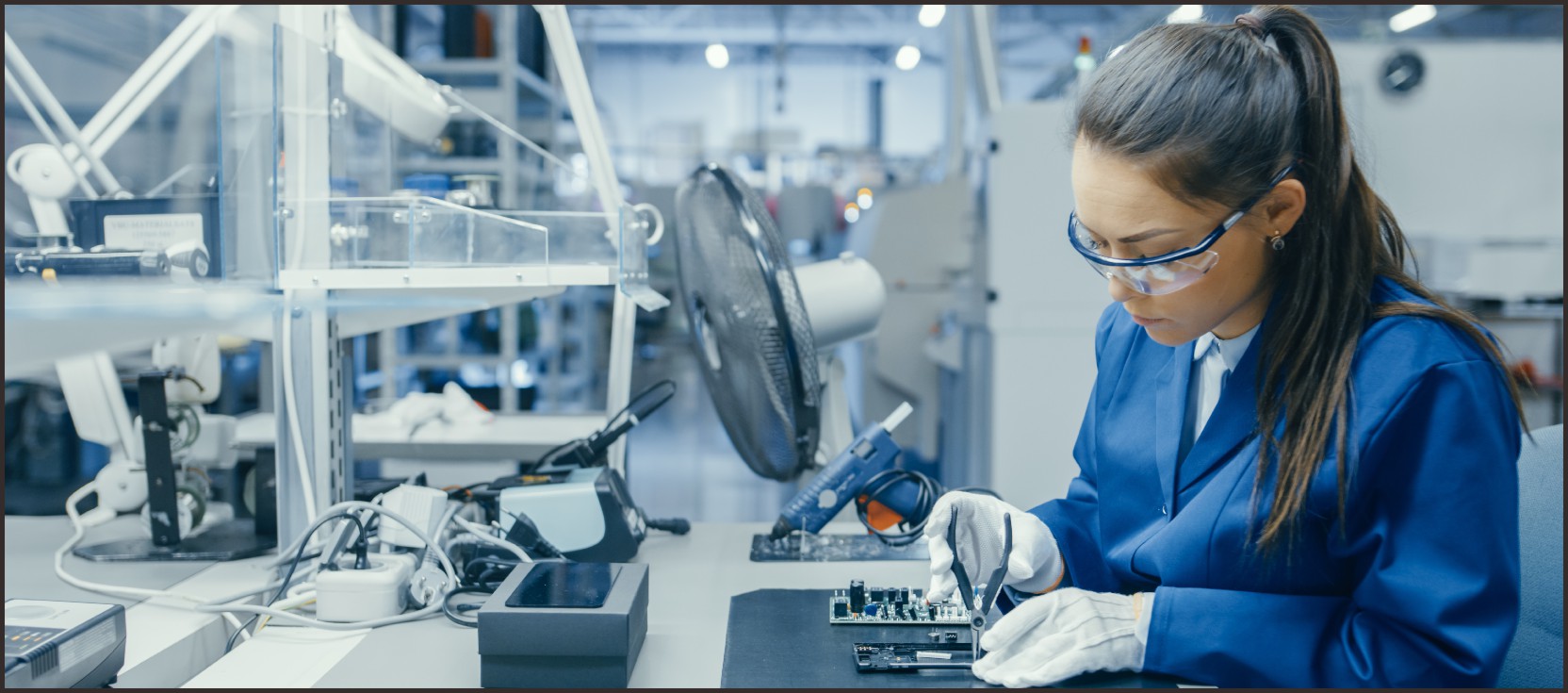 Un technicien en blouse de laboratoire bleue et lunettes de sécurité travaillant sur une carte électronique dans une installation de production high-tech, illustrant le processus de production personnalisé, de la planification et de la gestion à l'inspection de la qualité et à l'amélioration continue